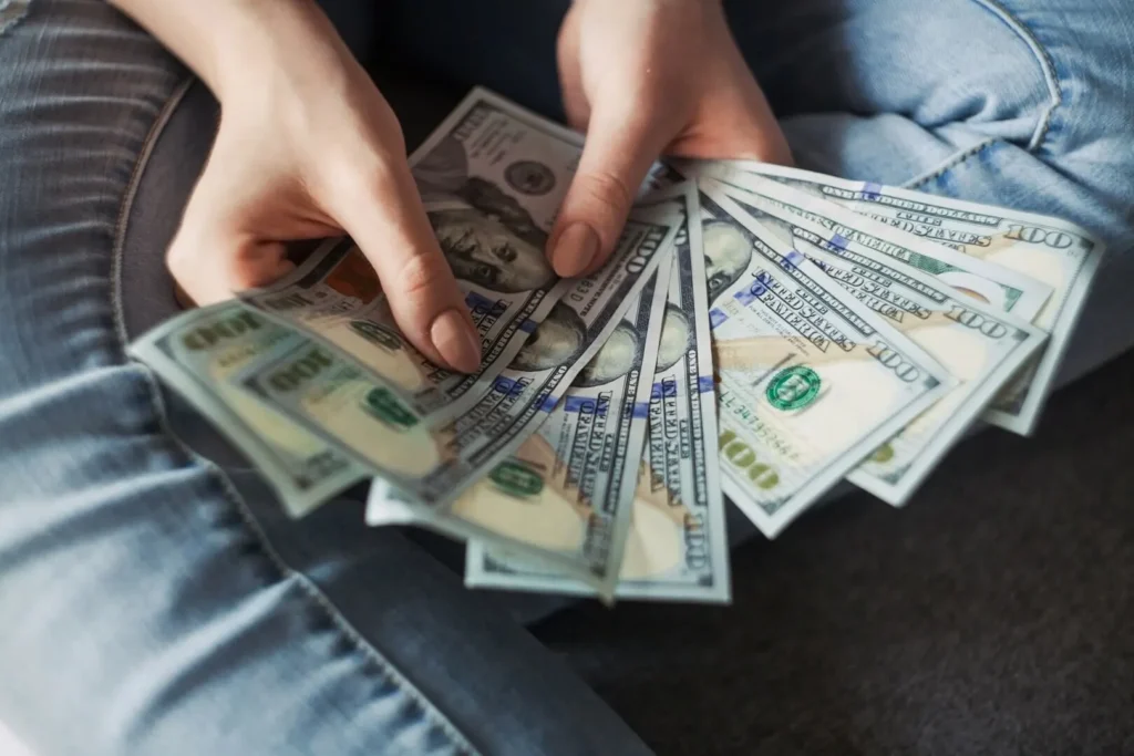 Person holding fanned out 100 US dollar bills on their jeans-clad lap